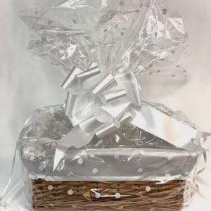 May include: A gift basket wrapped in clear cellophane with white polka dots. The basket is tied with a large white bow. The basket is made of woven brown wicker.