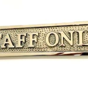 May include: A rectangular metallic sign with a polished finish featuring the text STAFF ONLY in raised capital letters. The background of the sign has a textured, dimpled pattern, and there are two circular screw holes on the far left and right.