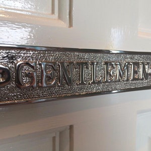 K&ouml;nnte beinhalten: Ein silbernes Metallschild mit dem Wort "GENTLEMEN" in Pr&auml;gung. Das Schild ist an einer wei&szlig;en T&uuml;r angebracht.