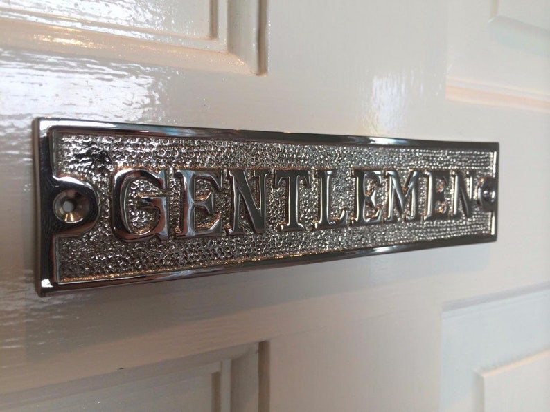 K&ouml;nnte beinhalten: Ein silbernes Metallschild mit dem Wort "GENTLEMEN" in Pr&auml;gung. Das Schild ist an einer wei&szlig;en T&uuml;r angebracht.