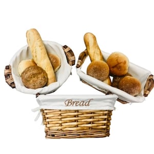May include: Three wicker bread baskets with white fabric liners. The baskets are filled with various breads, including baguettes and rolls. One basket has the word "Bread" embroidered on the liner.