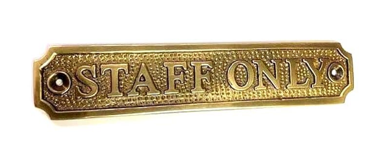 May include: A rectangular brass sign with a textured background and raised lettering that reads STAFF ONLY. The sign features a polished border and two circular mounting holes, one on each end, designed for attachment to a door or wall surface.