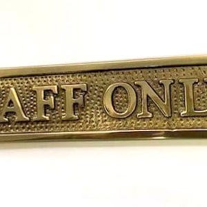 May include: A rectangular brass sign with rounded corners and a textured background. The sign features the raised, embossed text "STAFF ONLY" in capital letters. There is a small circular mounting hole on both the far left and far right sides.