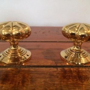 May include: A pair of polished brass door knobs featuring a rounded, fluted design with multiple vertical ridges. Each knob has a smooth, reflective metallic finish and is attached to a circular base plate with visible screw holes for mounting.