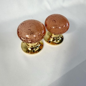 May include: A pair of round door knobs featuring translucent pink glass with internal air bubble details, mounted on polished gold-tone metal circular bases with visible screw holes for installation on a door surface.