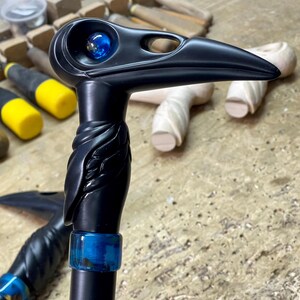 May include: A black walking stick with a carved raven head, blue glass eye, and a blue and green resin accent. The stick is on a wooden surface.