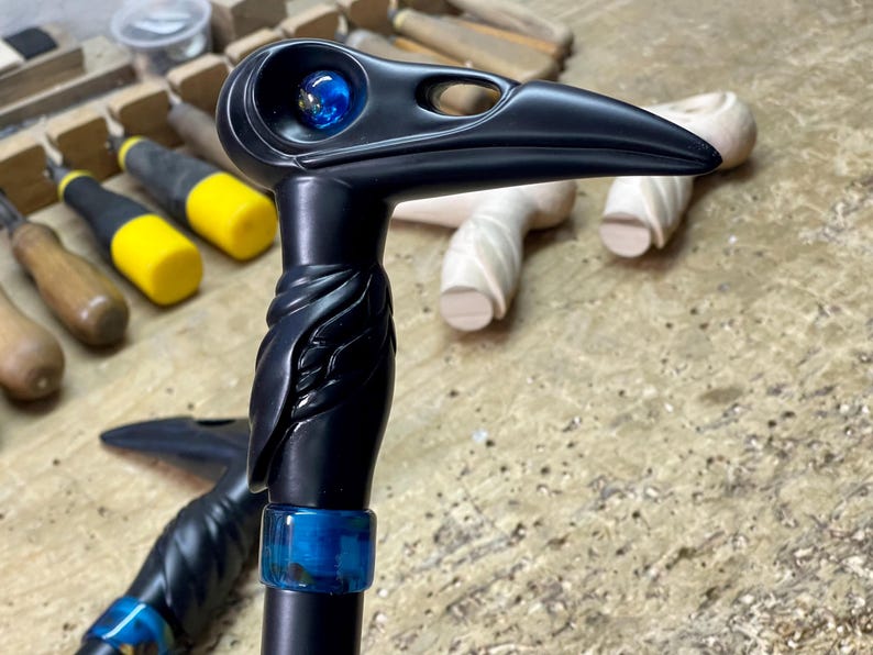 May include: A black walking stick with a carved raven head, blue glass eye, and a blue and green resin accent. The stick is on a wooden surface.