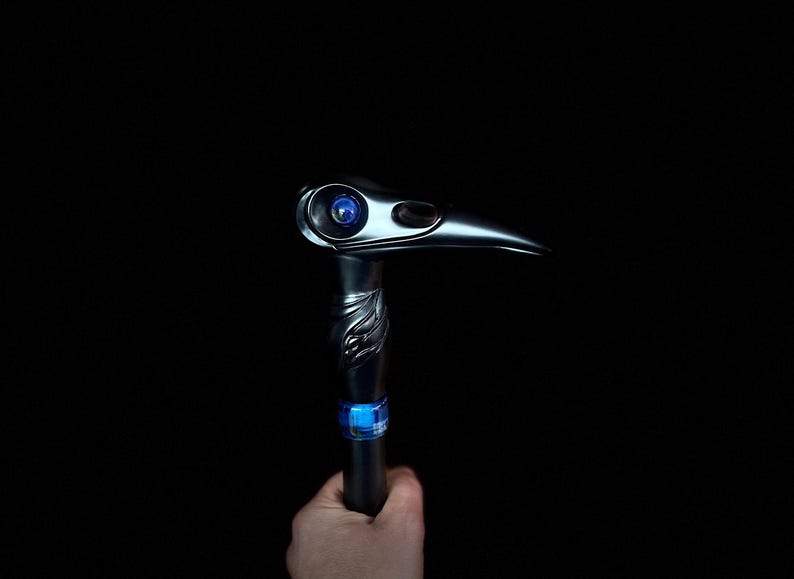 May include: A black walking stick with a bird's head design. The head has a blue eye and a long, curved beak. The stick has a blue and silver band around it.