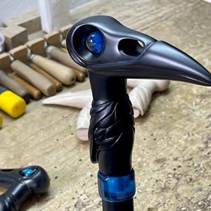 May include: A black walking stick with a carved bird head handle. The bird's eye is a blue glass marble. The stick is decorated with a blue band.