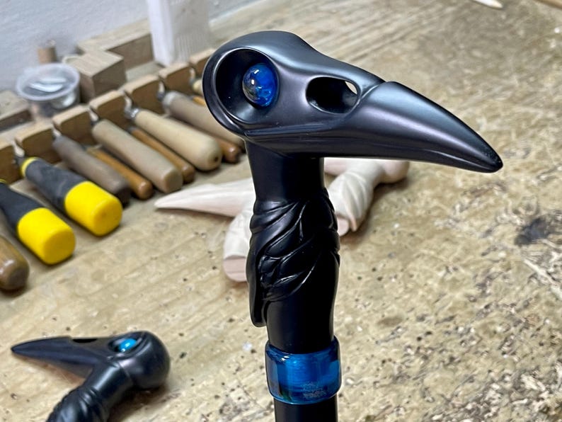 May include: A black walking stick with a carved bird head handle. The bird's eye is a blue glass marble. The stick is decorated with a blue band.