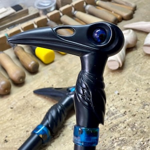 May include: A black walking stick handle carved in the shape of a bird's head with a blue eye. The handle is attached to a black walking stick shaft with a blue ring.