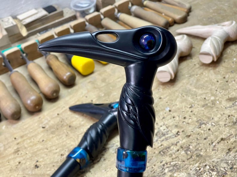 May include: A black walking stick handle carved in the shape of a bird's head with a blue eye. The handle is attached to a black walking stick shaft with a blue ring.