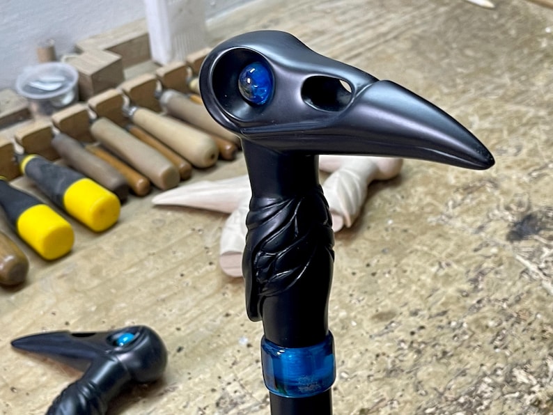 May include: A black walking stick with a carved bird head handle. The bird's eye is a blue glass marble. The stick is decorated with a blue band.