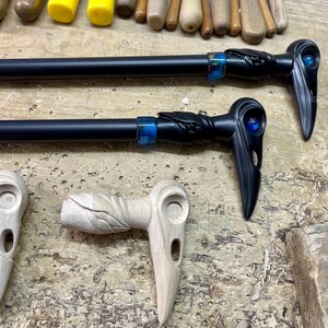 May include: Two black walking sticks with carved raven heads and blue glass eyes. The sticks are on a wooden surface with two unfinished wooden raven heads.