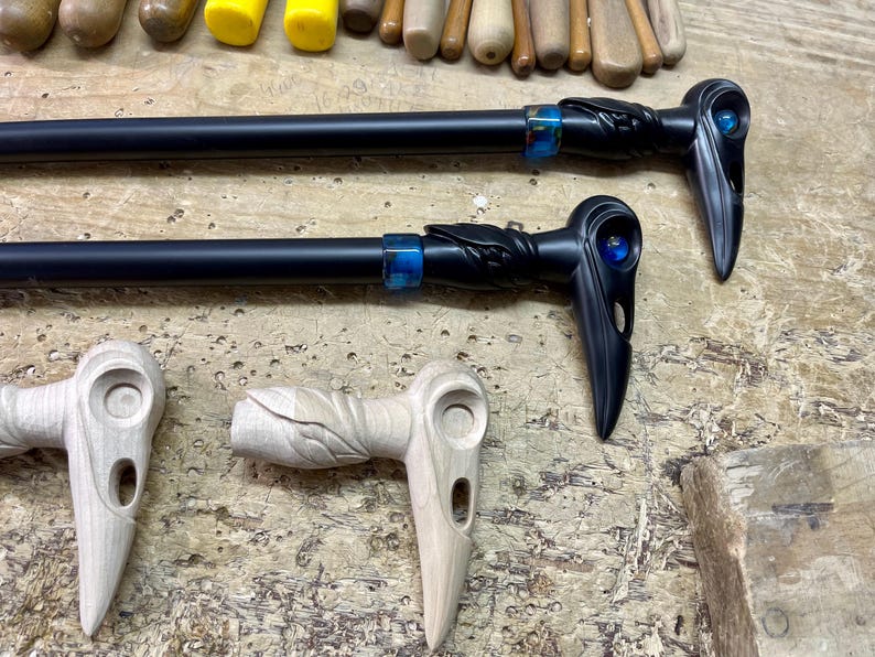 May include: Two black walking sticks with carved raven heads and blue glass eyes. The sticks are on a wooden surface with two unfinished wooden raven heads.