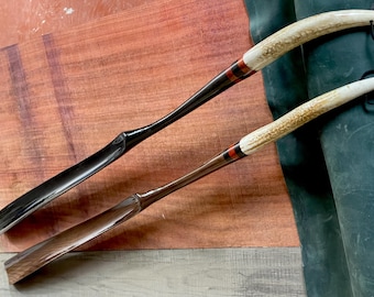 May include: Two wooden shoe horns with antler handles. The handles are decorated with red and black bands. The shoe horns are lying on a wooden surface.