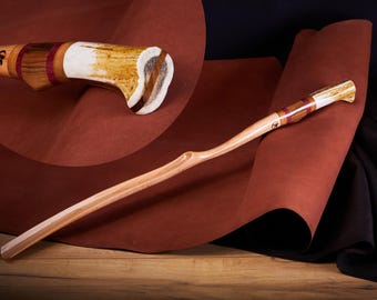 May include: A wooden shoehorn with a curved handle. The handle is made of wood with a white antler inlay and red and brown stripes. The shoehorn is on a brown leather surface.