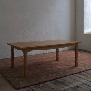 Op de afbeelding: Een lichtbruine houten eettafel met vier gedraaide poten. De tafel is rechthoekig en staat op een rood tapijt met patronen. De kamer heeft witte muren en een raam.