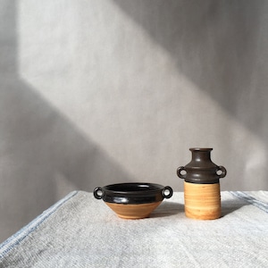 May include: A black and brown ceramic bowl with two handles and a black and brown ceramic vase with two handles. The bowl and vase are sitting on a white linen tablecloth.