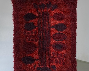 May include: A rectangular, shaggy rug in shades of red and burgundy, with a central design resembling a stylized tree. The rug is hanging from a metal rod with a cream-colored fringe at the top. The design features dark burgundy leaf-like shapes.