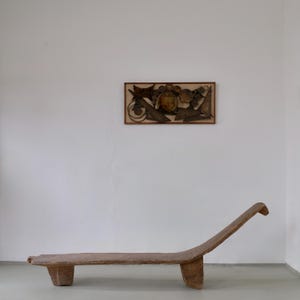 Op de afbeelding: Een houten chaise longue met een laag profiel, een gebogen, langwerpige zitting en twee stevige blokpoten. Boven de bank hangt een rechthoekig wandkunstwerk met een textuurrijke, abstracte compositie van donkerbruine geometrische vormen.