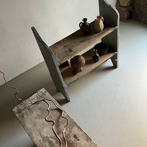 May include: A two-tiered wooden shelf unit painted in a light gray color. The shelf is made of reclaimed wood and has a rustic look. The shelf is decorated with a few small ceramic pots and a wooden sculpture.