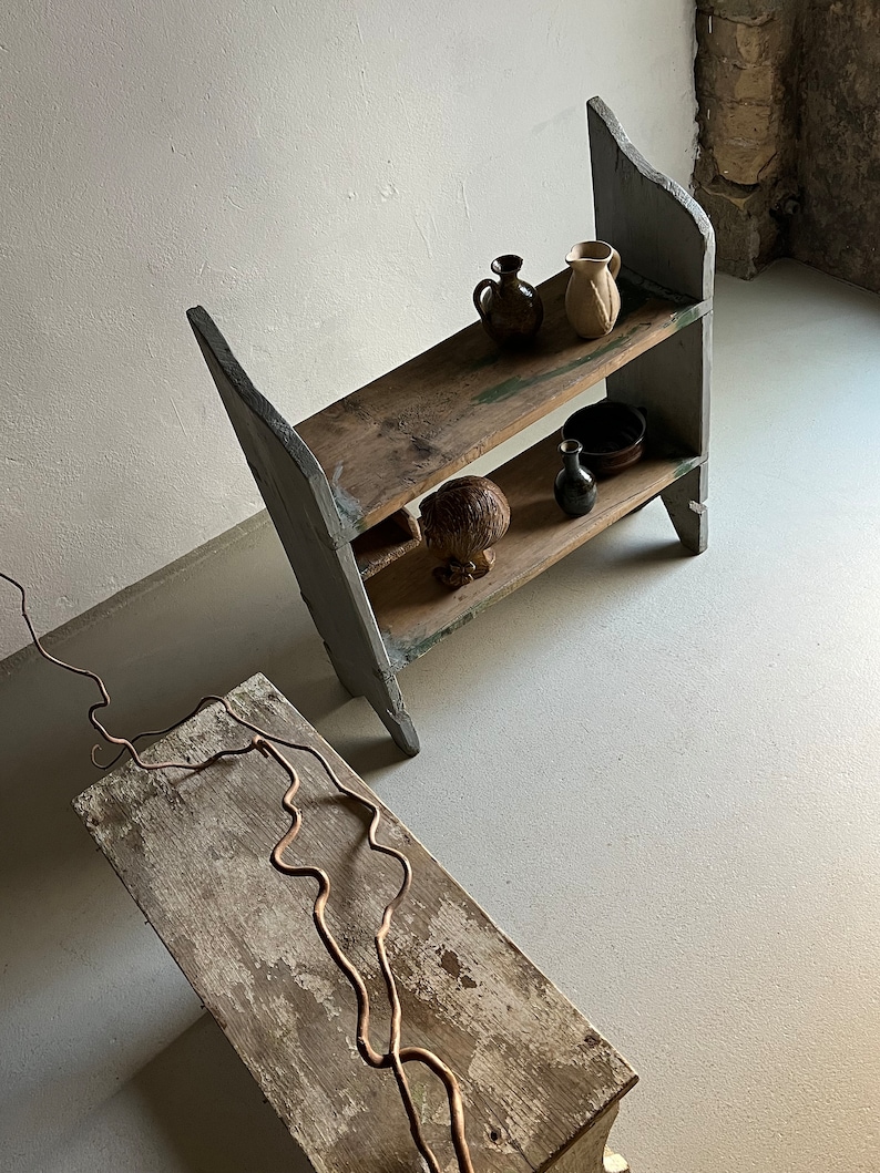 May include: A two-tiered wooden shelf unit painted in a light gray color. The shelf is made of reclaimed wood and has a rustic look. The shelf is decorated with a few small ceramic pots and a wooden sculpture.
