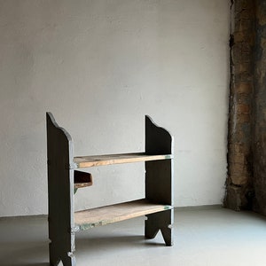 May include: A two-tiered wooden shelf unit with a rustic, distressed finish. The shelf is painted a dark green color and has two wooden shelves. The shelf is standing on a light gray concrete floor.