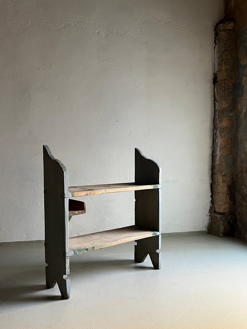 May include: A two-tiered wooden shelf unit with a rustic, distressed finish. The shelf is painted a dark green color and has two wooden shelves. The shelf is standing on a light gray concrete floor.