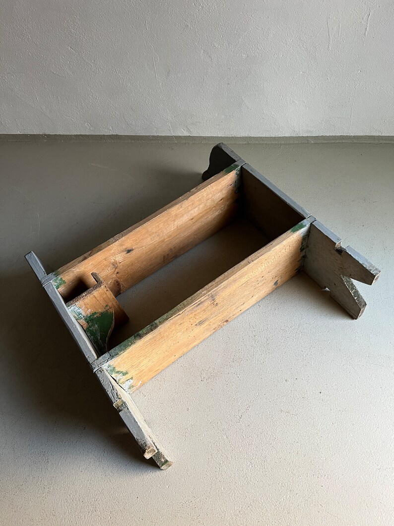 May include: A wooden stool with a gray and green paint finish. The stool has a simple design with two horizontal supports and a rectangular base.