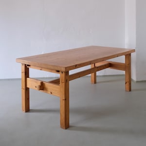 Pode incluir: Uma mesa retangular de madeira com acabamento castanho claro, onde são visíveis os veios e nós da madeira. A mesa é composta por quatro pernas quadradas robustas ligadas por vigas de suporte horizontais laterais e uma barra transversal central.