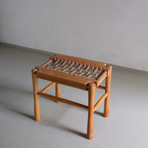 Puede incluir: Un taburete de madera con un asiento de cuerda trenzada. El taburete tiene un diseño sencillo con cuatro patas y un asiento rectangular.