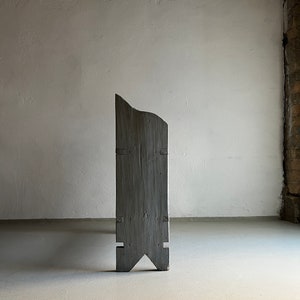 May include: A tall, gray wooden sculpture with a unique, asymmetrical shape. The sculpture has a pointed top and a wide base with two angled legs.