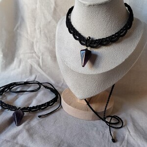 May include: A black macrame choker necklace with a dark purple, triangular pendant. The necklace is displayed on a mannequin head. A second necklace with a similar design is also visible.