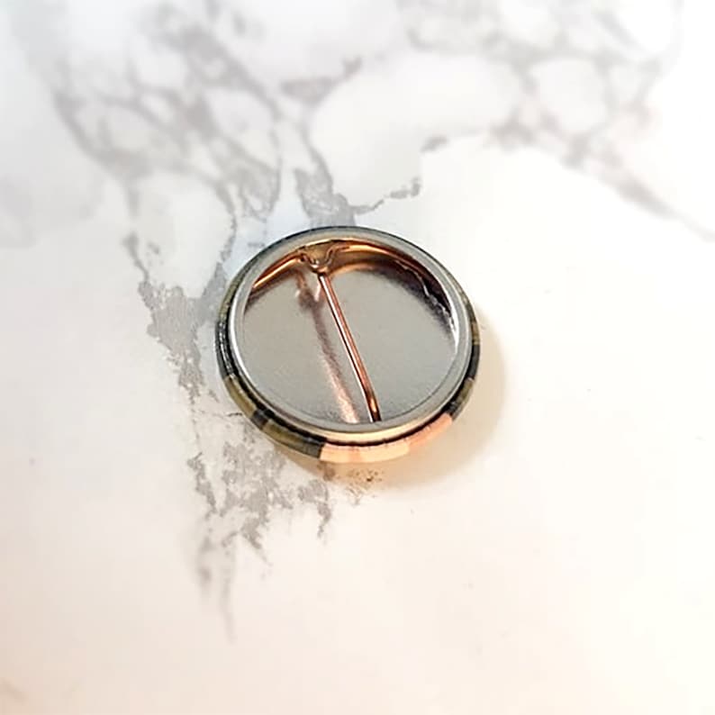 May include: The back of a round button with a metal pin. The button has a black, white, and green plaid design.