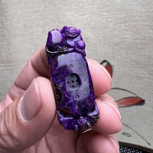 May include: Purple carved stone pendant with a detailed design of a mythical creature. The pendant has a small hole in the center.