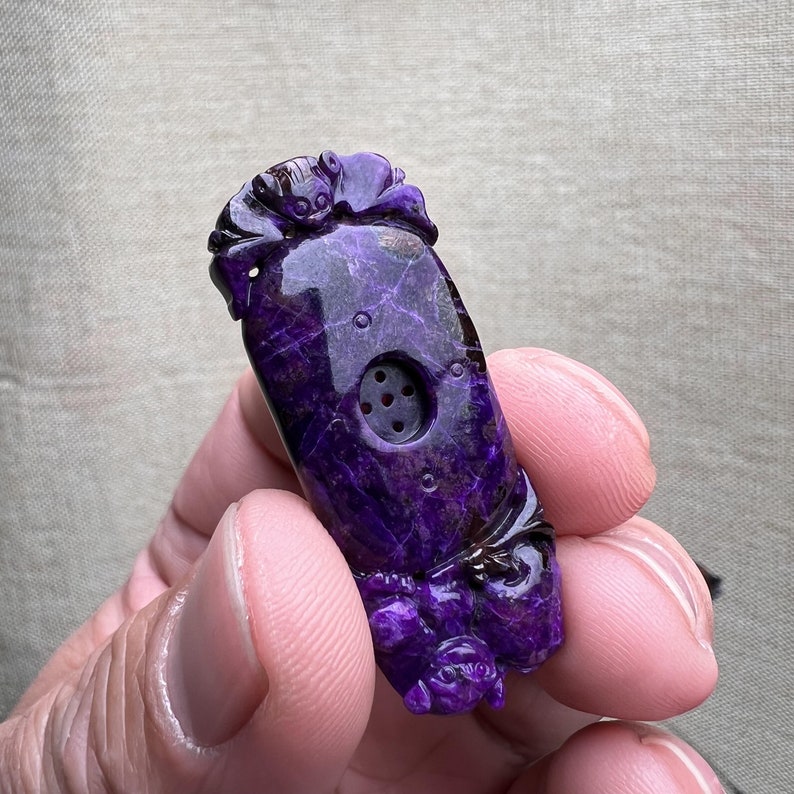 May include: Purple carved stone pendant with intricate details and a small hole in the center. The pendant is shaped like a gourd and has a textured surface.