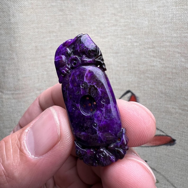 May include: A purple carved stone pendant with intricate details and a circular opening. The pendant is held in a hand.