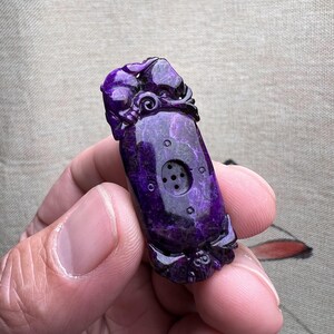 May include: Purple carved jade pendant with a detailed design and a small hole in the center.