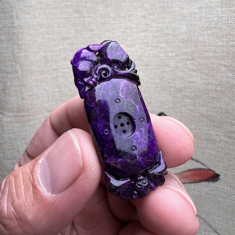 May include: Purple carved jade pendant with a detailed design and a small hole in the center.