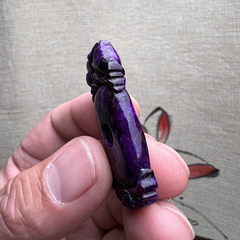 May include: A purple carved stone pendant with intricate details. The pendant is shaped like a figure with a round face and a long body. The stone has a glossy finish.