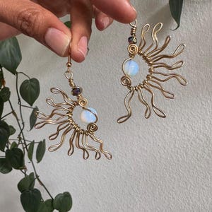 May include: A pair of handmade gold-colored wire earrings. Each earring features a sunburst design with swirling wire details and a central, round, opalescent bead. Additional small beads in purple and clear hues are also present.