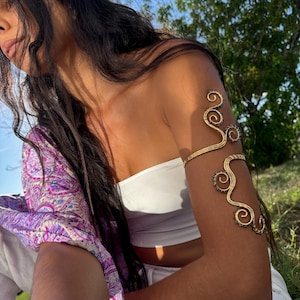 May include: Gold-toned arm cuff with a swirling design. The cuff features an upper arm band and a lower arm band connected by a decorative, curved design. Small, dark beads accent the design. The arm cuff is worn on a bare arm.