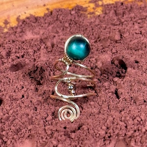 Puede incluir: Anillo artesanal de alambre dorado con una piedra preciosa redonda de color verde azulado. El anillo tiene un diseño en espiral con intrincados alambres y pequeños detalles de cuentas, sobre un fondo marrón rojizo.