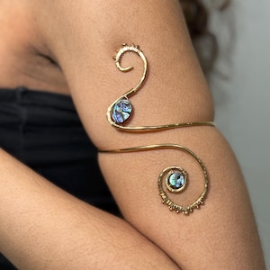 Könnte beinhalten: Ein goldfarbener Armreif mit einem wirbelnden Design. Der Armreif hat zwei schillernde Akzente aus Abalone-Muscheln. Der Armreif umschließt den Oberarm mit einer dekorativen Spirale oben und unten.