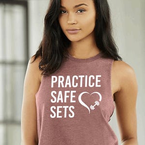Könnte beinhalten: Ein weinrotes ärmelloses Workout-Top mit dem weißen Text "PRACTICE SAFE SETS" sowie einem Herz- und Hantel-Grafik. Das Top ist aus einem weichen Material gefertigt.