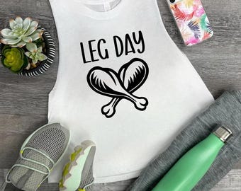 May include: A white sleeveless tank top featuring the black text LEG DAY above a graphic of two crossed chicken legs. The shirt is displayed alongside a succulent in a pot, a phone with a colourful leaf pattern, grey trainers, and a light green bottle.