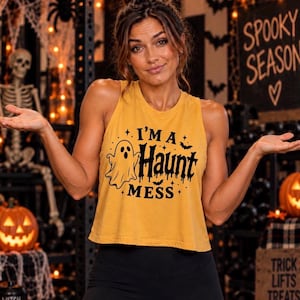 May include: A mustard yellow sleeveless crop top featuring a black graphic print of a ghost and bats with the text "I'M A HAUNT MESS" in a stylized font. The garment has a round neckline and a relaxed fit, constructed from a cotton-blend fabric.