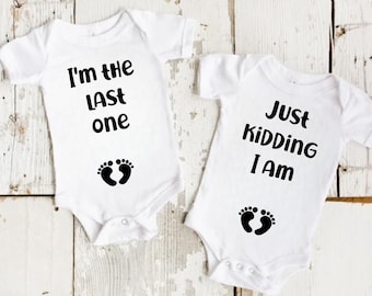 May include: Two white baby onesies with black text and footprints. One says "I'm the LAST one" and the other says "Just Kidding I Am". The onesies are on a light wood background.
