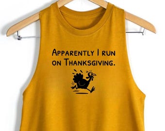 May include: A mustard yellow sleeveless crop top featuring the black text "Apparently I run on Thanksgiving." and a black graphic illustration of a running turkey centred on the front. The garment is displayed on a wooden clothes hanger.